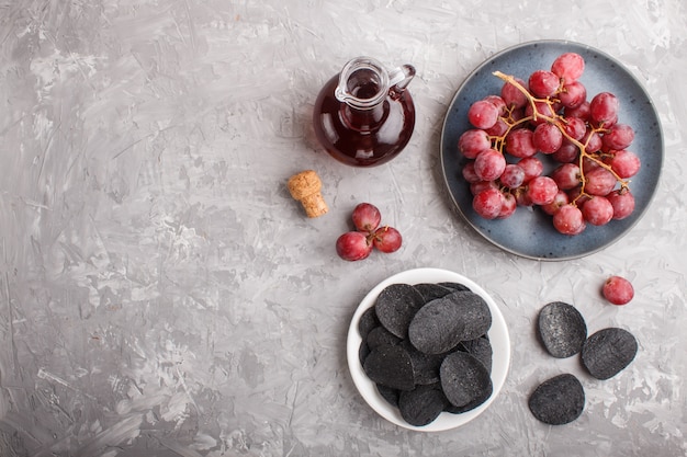 Black potato chips with charcoal, balsamic vinegar in glass, red grapes on a blue ceramic plate . Top view.