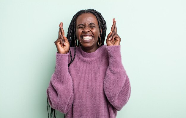 Black pretty woman smiling and anxiously crossing both fingers, feeling worried and wishing or hoping for good luck