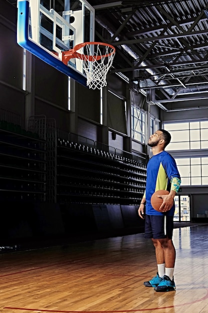 Black professional basketball player holds a ball over the hoop in a game hall.