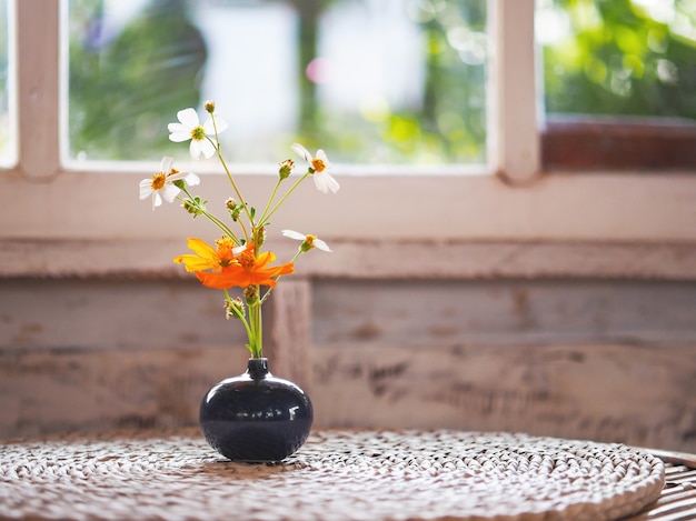 Black round vase with flower bouquet on vintage wooden table nearby glass window in living room at sunset.