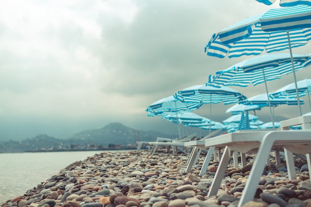 Black sea and pebble beach with beach umbrellas and deck chairs against mountains. Sea vacation and relaxing time