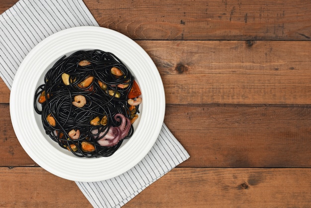 Black seafood pasta in white plate over linen towel on rustic wood background with copy space. 