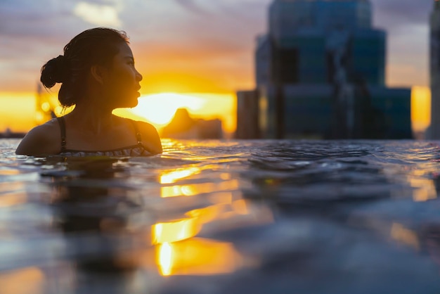 Siluetta nera della donna asiatica spruzzata d'acqua durante le vacanze estive rilassante nella piscina a sfioro con vista tramonto sul mare blu con grattacieli del centro urbano del centro urbano sano stile di vita di felicità