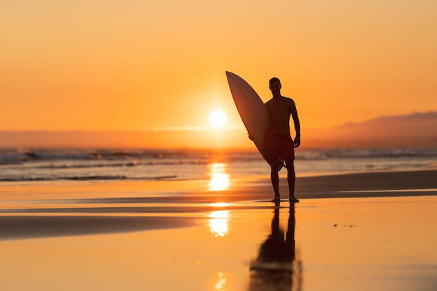 Черный силуэт привлекательного мужчины на берегу с доской для серфинга на оранжевом закате