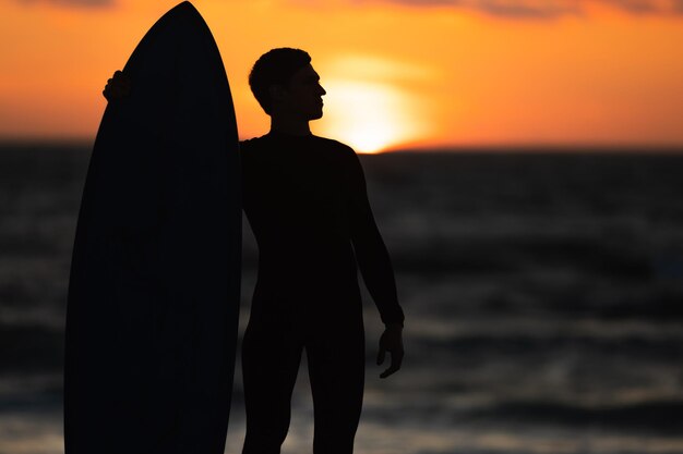Black silhouette of a man in wetsuit standing on the seashore holding a surfboard at bright orange s