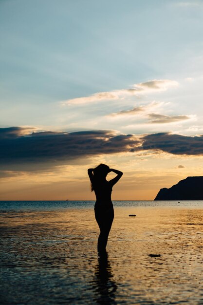 Black silhouette of woman posing in shallow water of ocean in tropics in sunset time