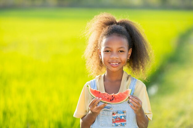 Black skinned cute little girl eating watermelon outdoors green rice field backdrop African child eating watermelon