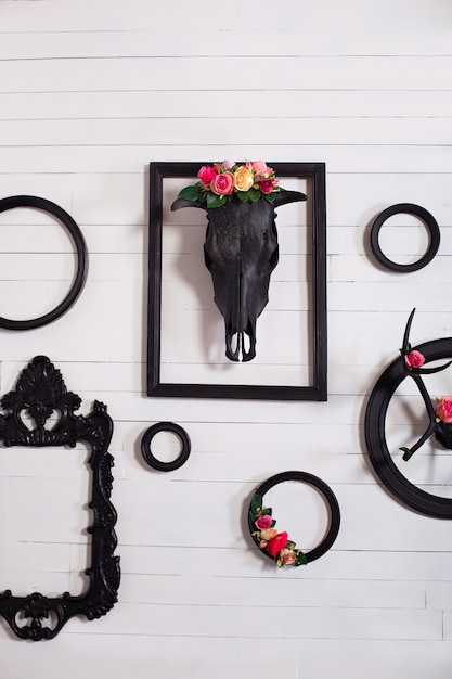 Black skull of a deer and horns on a wooden white wall with empty frames for paintings. The concept of decorating a wooden white wall in the Gothic style in the living room. Decor, vintage, modern