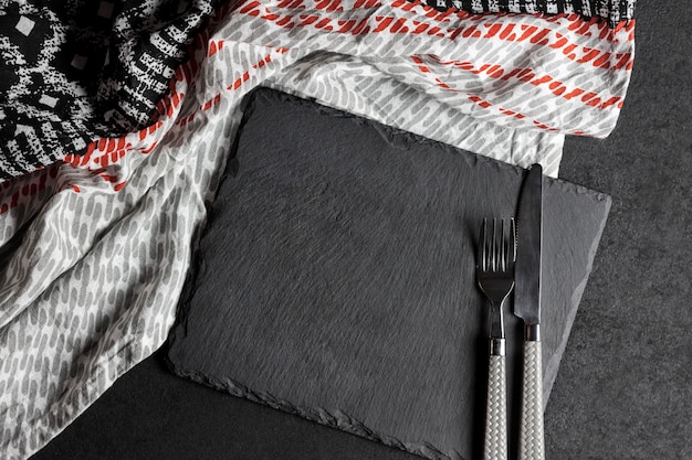 Black slate plate with fork and knife on black background and tablecloth. Table setting.