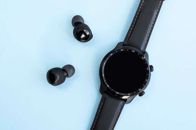Black smart watch and wireless headphones on blue background Blank screen copy space