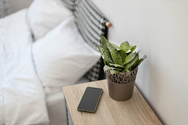 Black smartphone on the nightstand. Green plant in a flower pot on the table in the bedroom