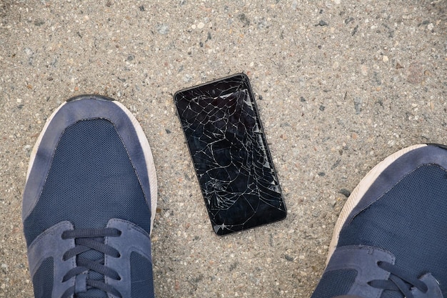 아스팔트 위에 있는 사람의 발 근처에 깨진 디스플레이가 있는 검은색 스마트폰
