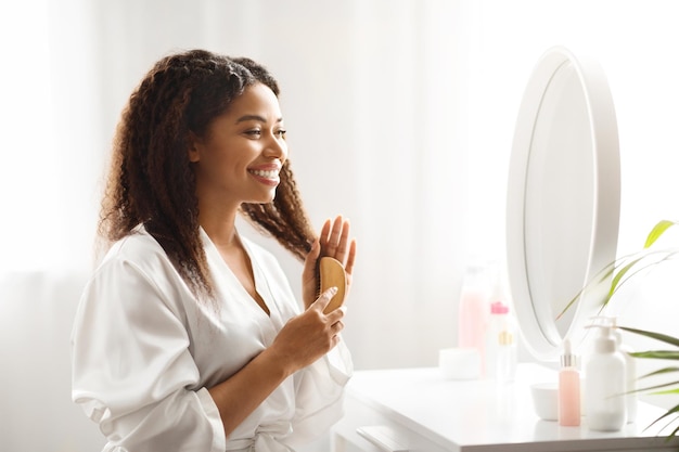 Black smiling female combing her beautiful hair with bamboo brush at home