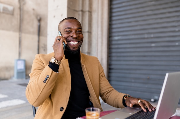 Черный улыбающийся человек разговаривает по телефону, сидя в кафе с помощью портативного компьютера.