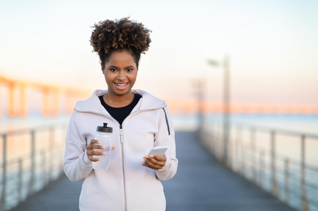 Black smiling woman in sportswear with smartphone and fitness bottle walking outdoors