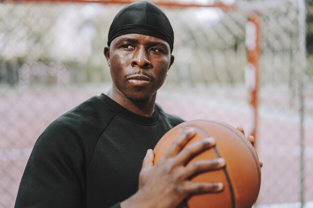 Black Spanish male in black casual clothes and do-rag holding a basketball ball on an outdoor court