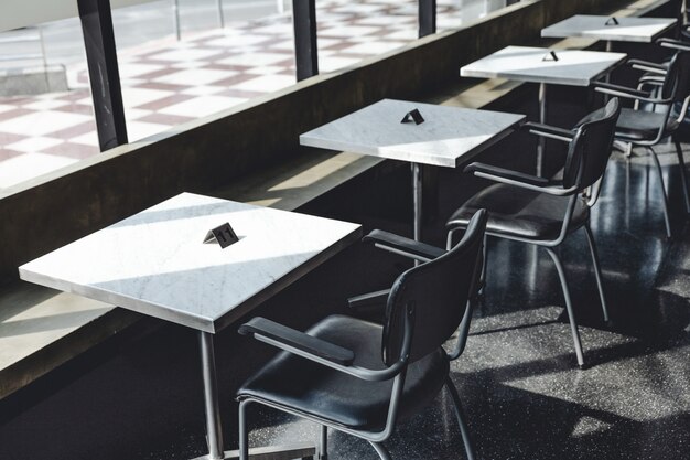 Black steel chairs and square white granite top table near glass window with direct sunlight. 