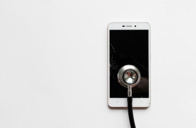 Черный стетоскоп на сломанный мобильный смартфон после падения, вид сверху, на белом фоне. Копировать пространство