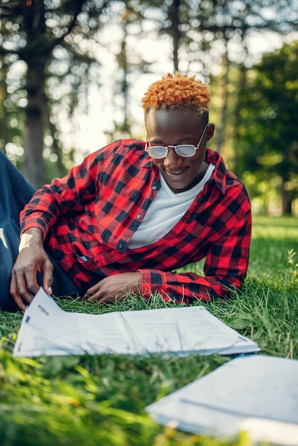 Black student in glasses reading book on the grass in summer park. 
