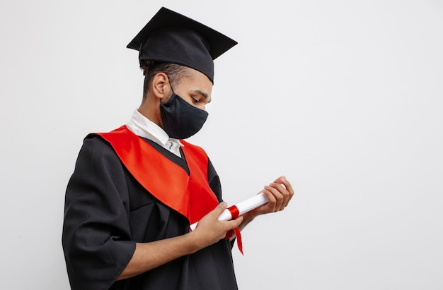 A Black student in graduate gown and square cap and a black face mask who is happy to finish his studies look at his the longawaited diploma Class of 2021