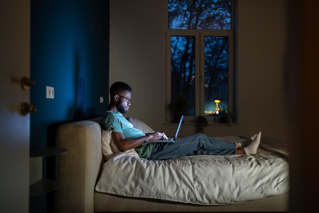 Photo black student guy lying in bed with laptop in evening studying until late hours