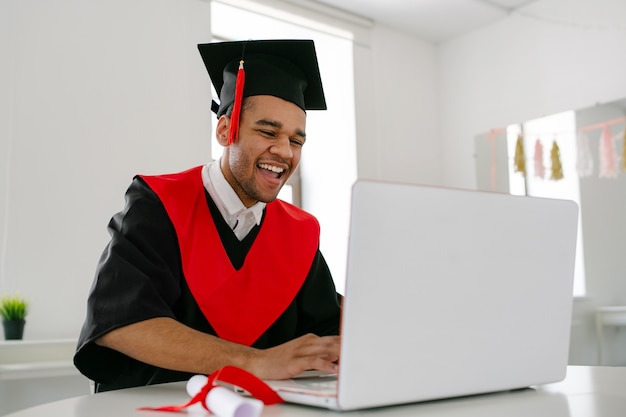 A Black student sits at a laptop in a gown and square cap and communicates with fellow graduates Virtual graduation and convocation ceremony Grag in light room copy space