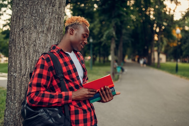 Черный студент с книгой и рюкзаком, стоящим возле дерева в летнем парке. Подросток учится на открытом воздухе и обедает