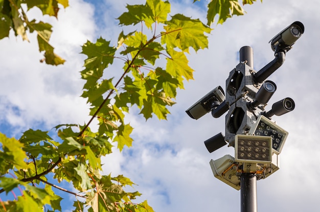 Videocamere di sorveglianza nere e lampioni bianchi su una colonna contro il cielo e le foglie verdi dell'albero di acero