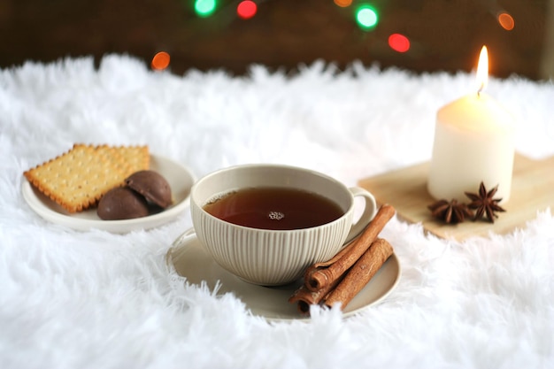 Фото Черный чай в кружке стоит на пушистом пледе за доской с новогодней гирляндой рядом стоит свеча конфеты палочки корицы и анис лежат