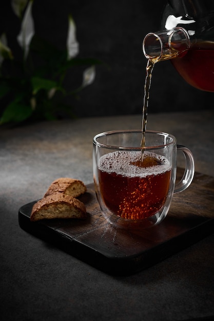 Black tea is poured from the teapot into transparent cup with bubbles nearby cookies on cutting