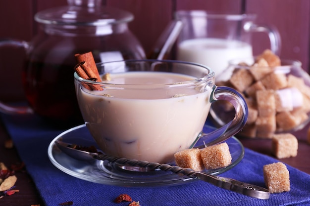 Black tea with milk and lump sugar and spices in glassware on color wooden planks background