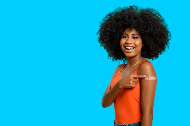 Black teen girl smile and points to her arm with vaccine sticker, she does not wear face shield, isolated on gray background.