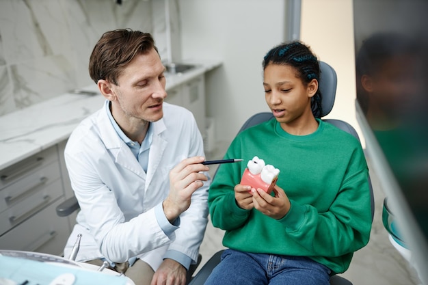 Black teenage girl sitting in dental chair and holding tooth model