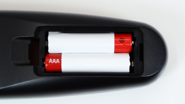 Black TV remote control with AAA alkaline batteries in red and white on a white background. Battery replacement, spare parts. Remote control battery compartment close-up.