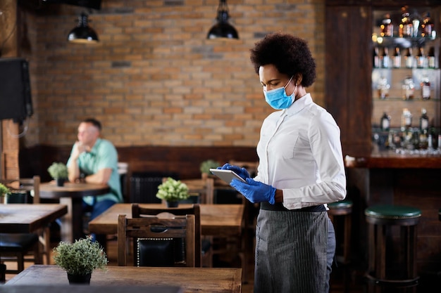 Black waitress with protective face mask and gloves working on touchpad in a cafe