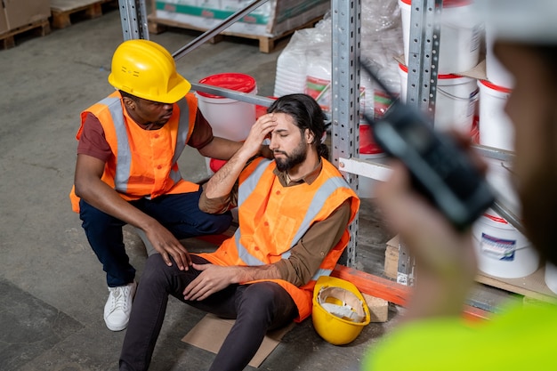 Black warehouse employee in orange vest holding shoulder of colleague and checking his condition after incident