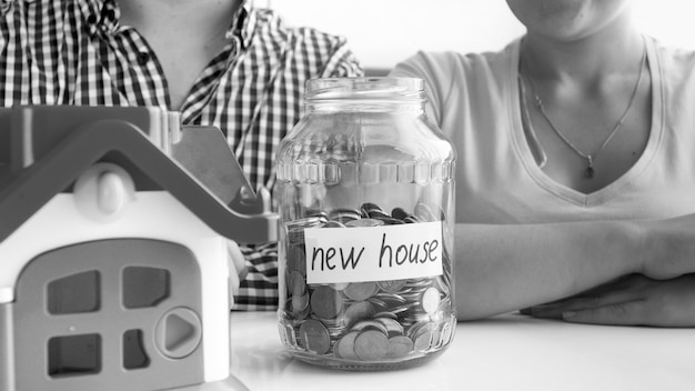 Black and white closeup view of young couple sitting at the white table on which is located small house and ,full of coins, jar with the inscription new house