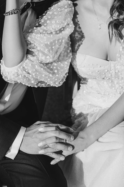 Black and white crop photo of bride and groom's hands in wedding dress. Unrecognizable persons.