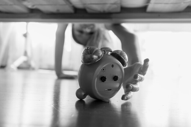 Black and white image of beautiful girl reaching for alarm clock under the bed