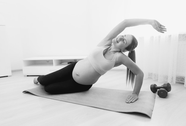 Black and white image of beautiful pregnant woman stretching on fitness mat at home