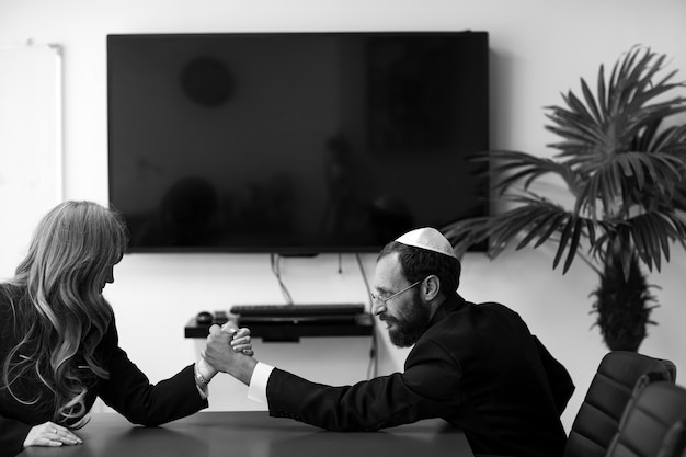 Photo black and white image of israeli business partners competing in arm wrestling in office. jewish man in yarmulke and woman with blond hair, tense faces compete armwrestling. feminism, gender equality