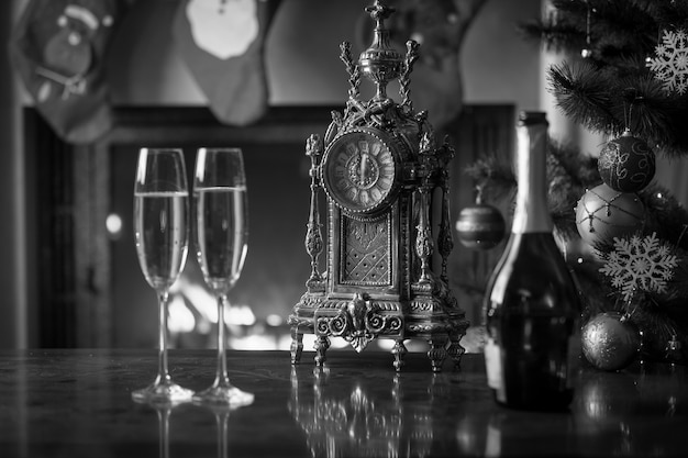 Black and white image of two glasses of champagne on table next to old clock show 12 o'clock. Monochrome Christmas background