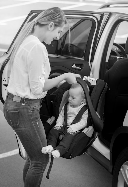 Black and white image of young mother putting car seat with her baby boy in the car