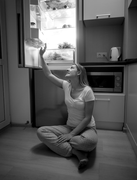 Black and white image of young woman sitting on floor and reaching for food from open refrigerator at late night