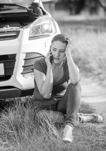 Black and white image of young woman sitting on ground and leaning on broken car