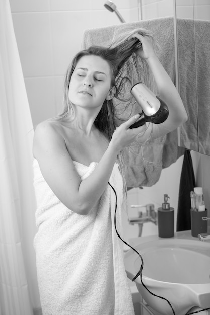 Black and white image of young woman in with hairdryer at bathroom mirror