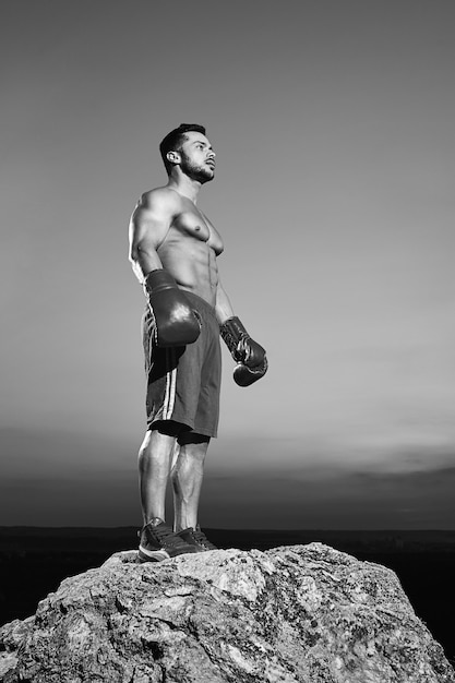 Черно-белый снимок под низким углом красивого молодого сильного мускулистого спортивного мужчины в боксерских перчатках, задумчиво глядя в сторону после тренировки на открытом воздухе по боксу спортивной мотивации Copyspace.