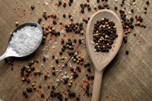 Black and white pepper peas sea salt cloves spices in a spoon\
on a wooden background