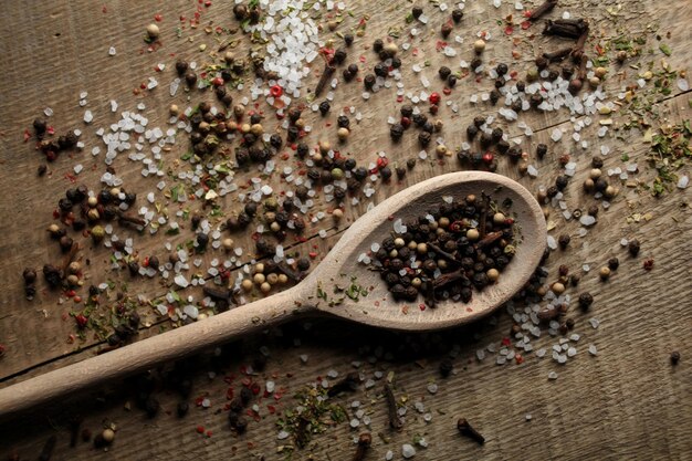 Black and white pepper peas sea salt cloves spices in a spoon\
on a wooden background