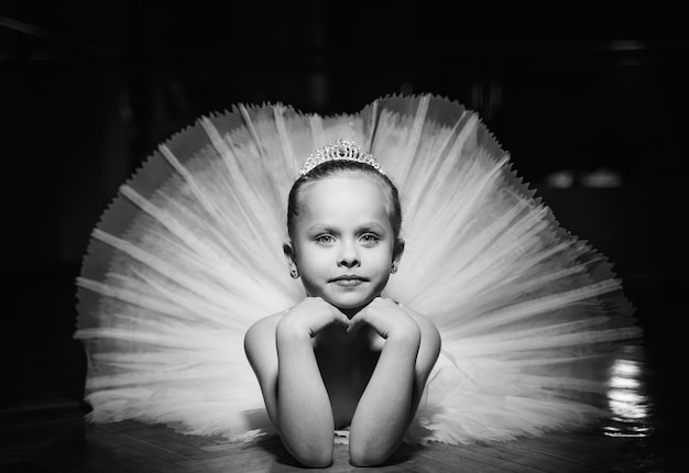 Black and white photo of a cute smiling ballerina in white tutu and a crown laying on the floor with hands under the chin. 